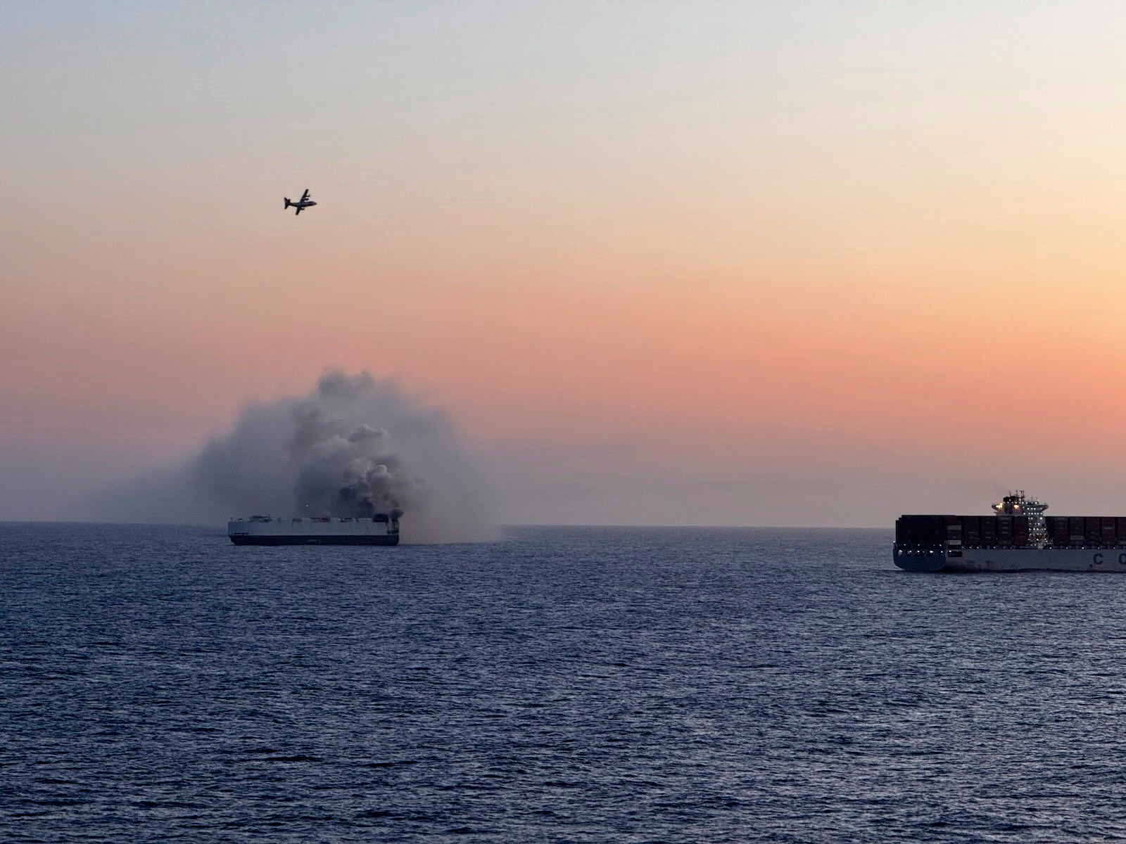 Cargo ship fire with smoke billowing, and a plane overhead.