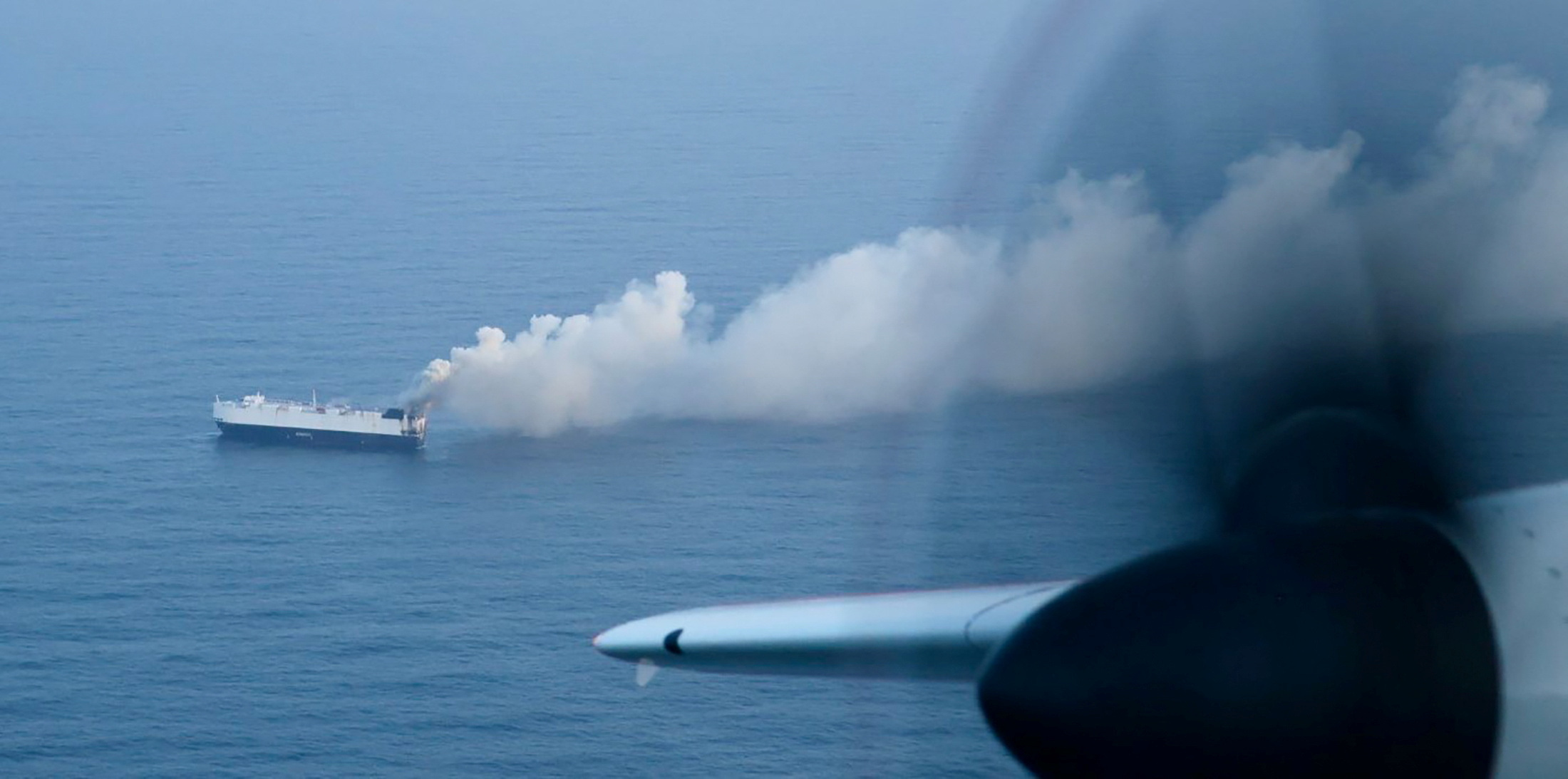 Aerial view of a cargo ship on fire in the ocean, with smoke billowing from it.