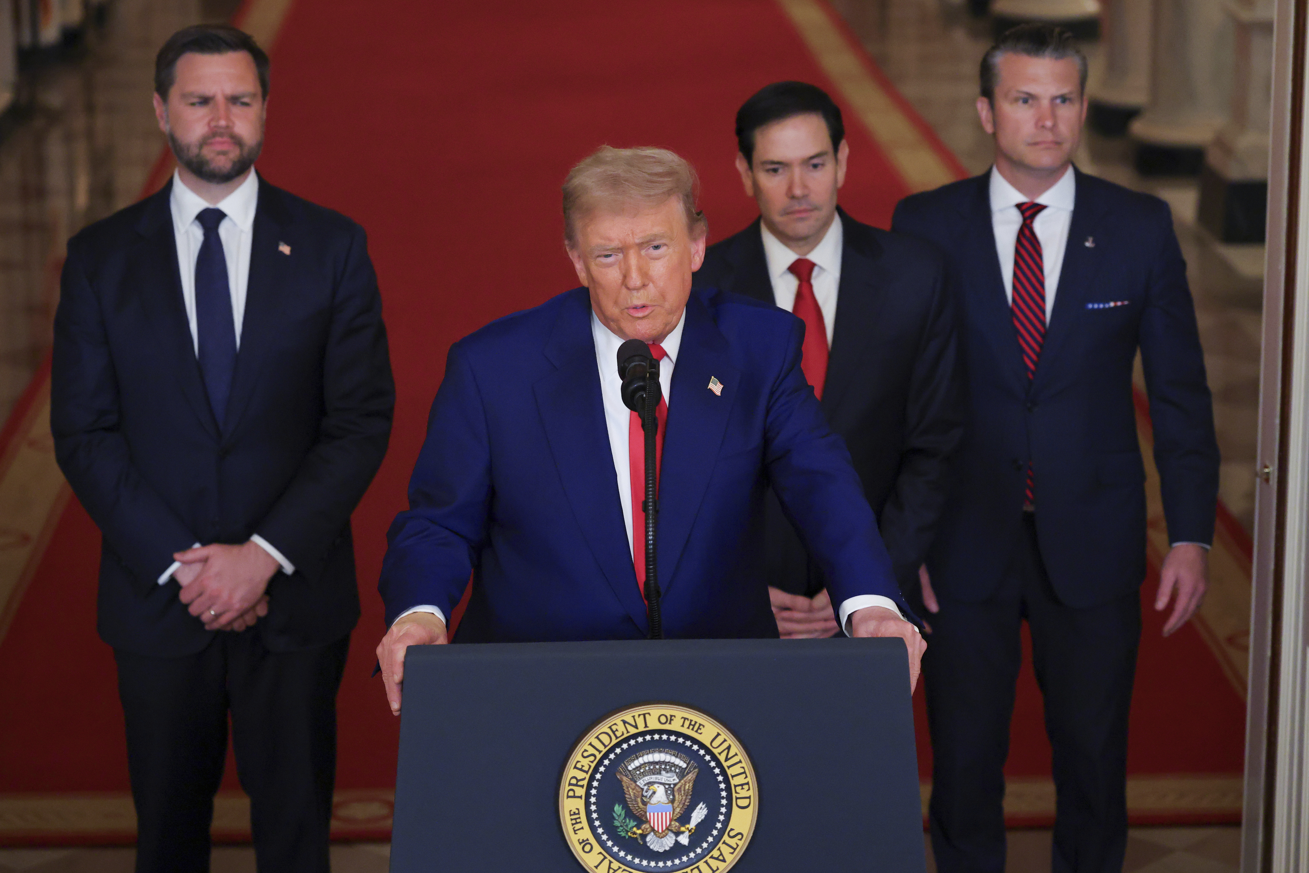 President Trump speaking at a podium with three men in suits standing behind him.
