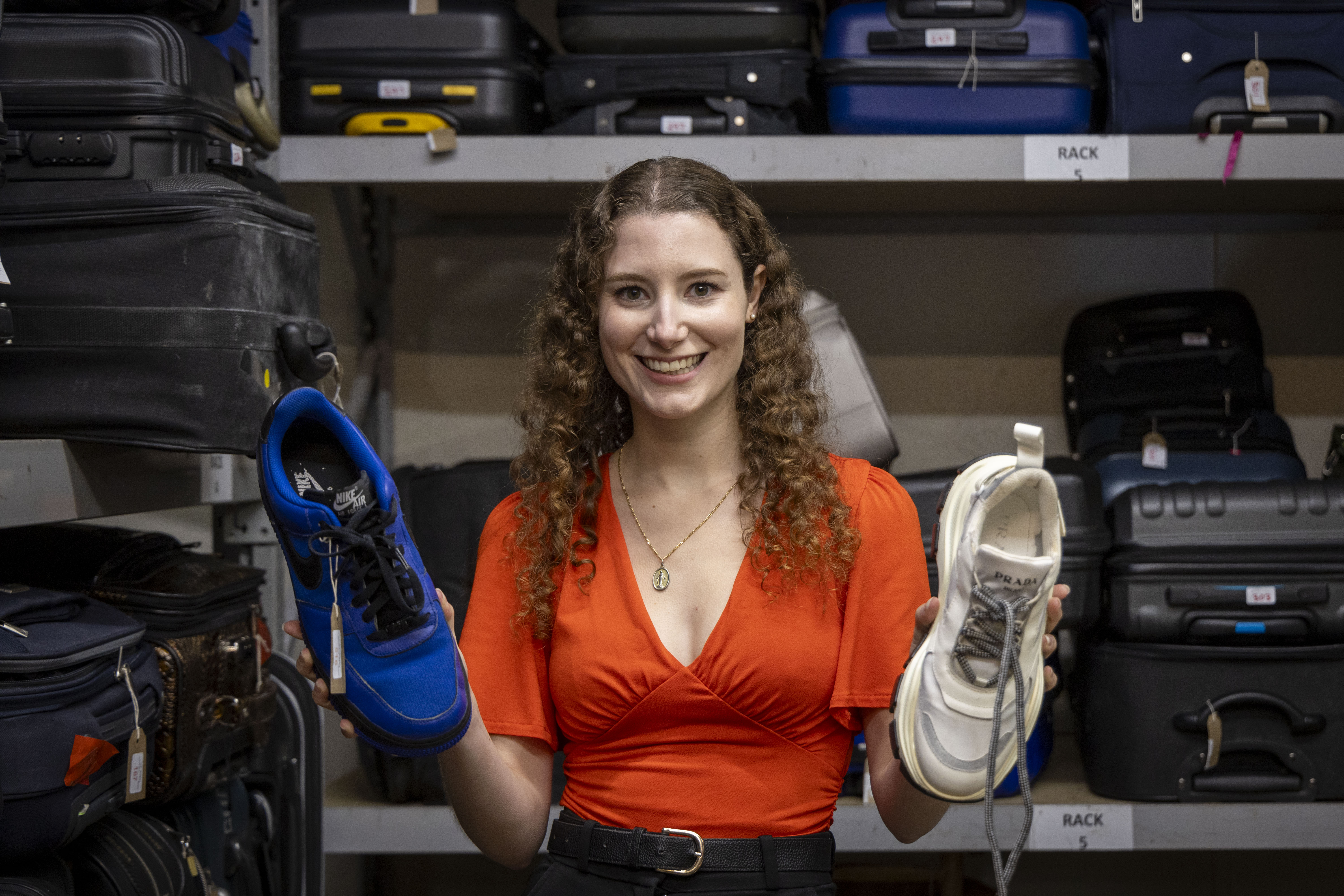 Woman holding up a blue Nike Air Force 1 and a white Prada sneaker in front of shelves of lost luggage.