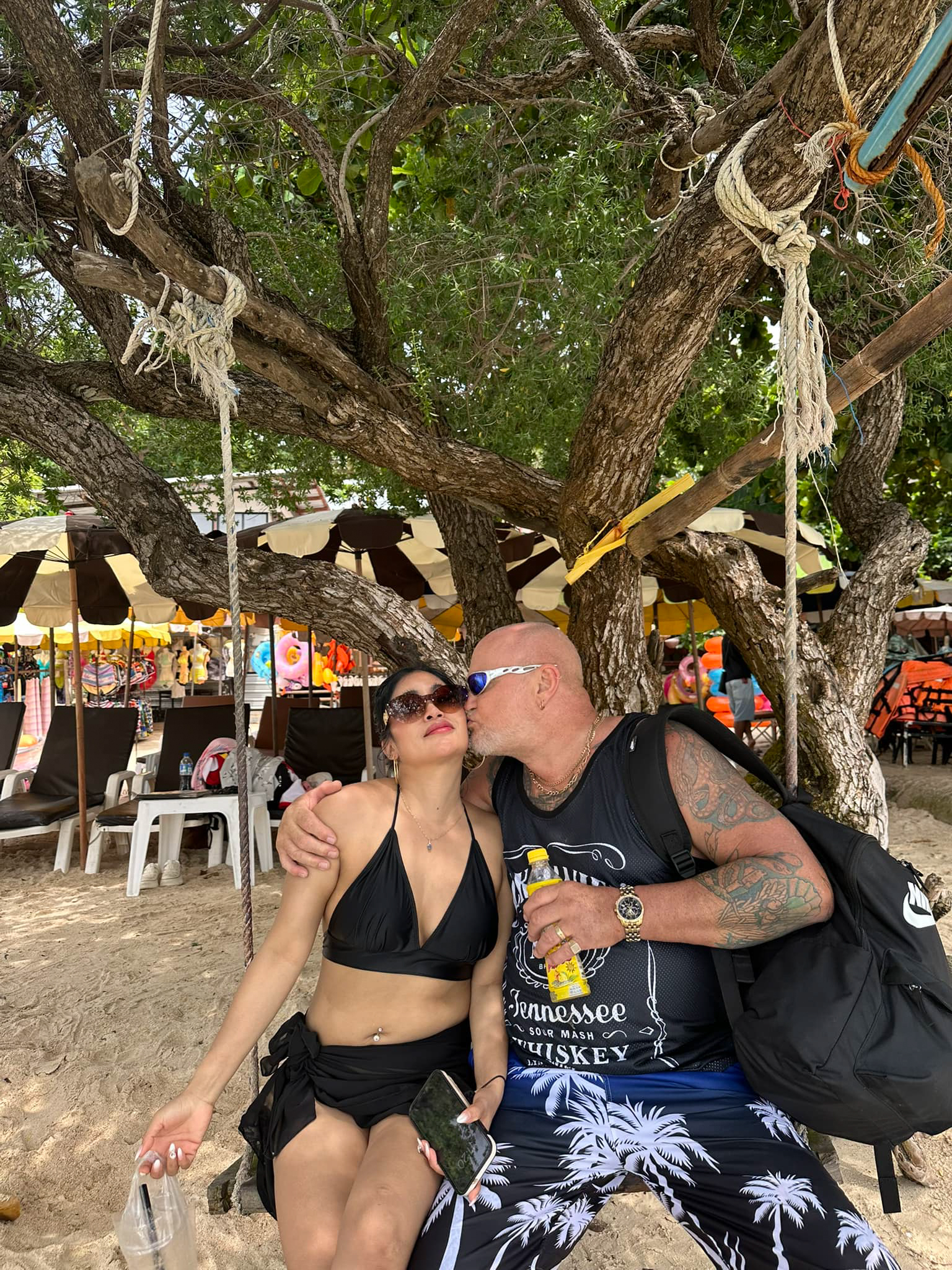 Photo of a man and woman kissing on a tree swing on a beach.